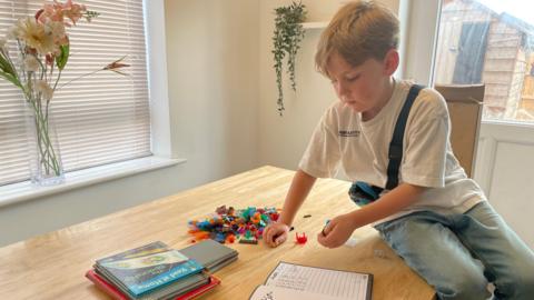 Austin sat on a wooden table which has books and LEGO blocks in a pile.