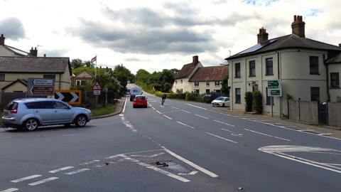 A crossroads at Earl Stonham in Suffolk. There are houses on either side. The A140 is running through the middle of the picture. Cars are waiting to join it from both sides of the crossroads.