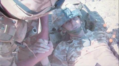 Two soldiers in desert camo army gear. One solider who's face is not in shot is holding the hand of a another solider Johno Lee lying on the floor who has been injured in IED blast. His head is resting on his backpack.