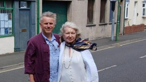 Nigel and Elaine Humes standing in front of one of the buildings where the new association will be. They both have white hair, which is swept by the wind. Nigel is wearing a burgundy coat over a blue and white striped shirt. Elaine is wearing a white shit and blouse. She has a satin navy blue scarf with a yellow print.