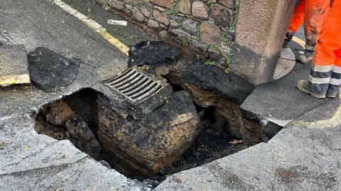 A picture of the sinkhole under the road. A hole in the road surface can be seen with workers attending the scene.