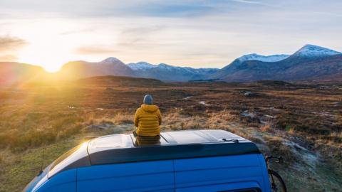 A man sits looking out at a beautiful and vast British landscape with the sun setting in the distance behind mountains.