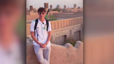 Tommy Lamb, 18, stands against a wall overlooking a town in Iran. He wears a white t-shirt, grey trousers and dark sunglasses. He has curly, brown hair.