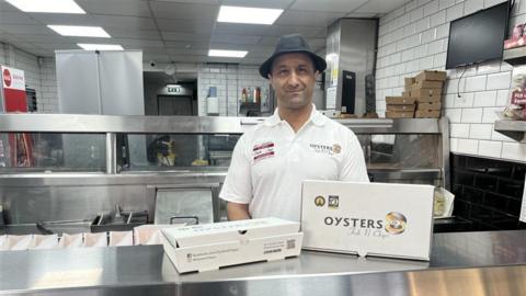 Gav Singh, a man wearing a black hat and a white shirt with "Oysters" written on the right side. He is stood in a fish and chip shop behind a metal counter with a cardboard container reading "Oysters" on the front.