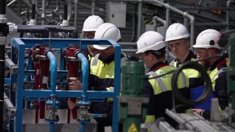 A number of people stood together working on an item in a warehouse. They are all wearing hard hats, glasses and a hi-vis.
