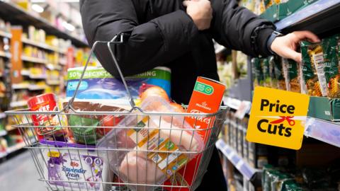 A shopping basket full of products being carried on a shopper's arm in a supermarket aisle. There is a sign on the shelf that says price Cut.