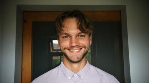 A man in a light purple shirt smiling in an internal doorway, grey walls to either side. He has a neat beard, a long brown fringe and blue eyes.