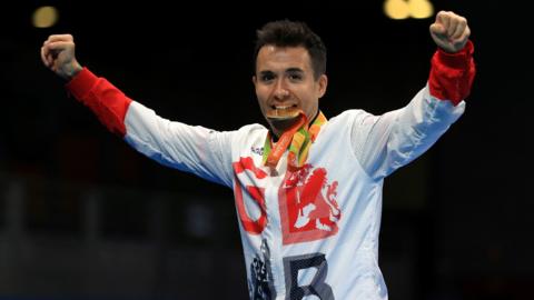 Will Bayley, wearing a GB sports shirt as he bites a gold medal. Both his arms are stretched out to the side in victory.