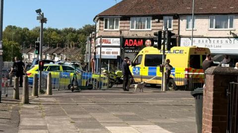 A yellow police van, a police motorbike and a police car are parked on the side of a road by a row of shops, behind a section of police tape. Passers by are stood looking on. 