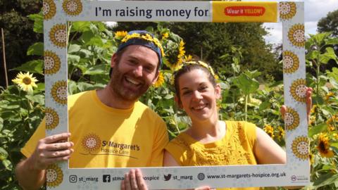 A man and women holding up social media frame with yellow T-shirts on trying to raise money for St Margets Hospice in front of sun flowers.