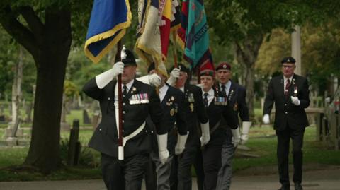 Five standard bearers, fully dressed in military uniforms, march in a cemetery in Hull. 
