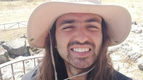 A bearded man with a cream hat on, with long brown hair stood infront of rocks