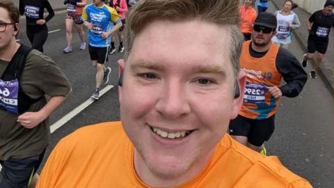 A close up shot of a man smiling in orange running gear mid-race. In the background there are other runners with number badges on.