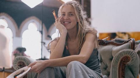 A woman with long blonde hair, wearing grey trousers and a vest top, sits on an armchair holding a plate palette and a brush