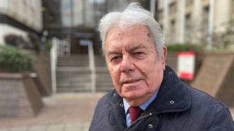 Mike Mackrory, a man in his 70s, is smiling at the camera with an out of focus council building behind him.