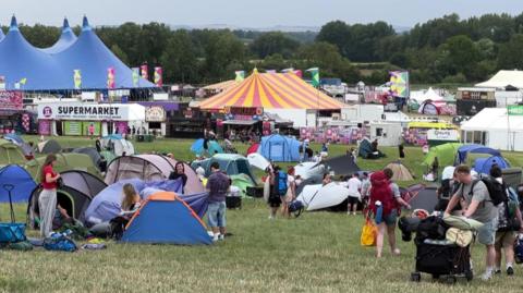 A wide view of a festival field with people pitching tents in the foreground, larger circus-style tents and flags behind and trees and countryside in teh distance.