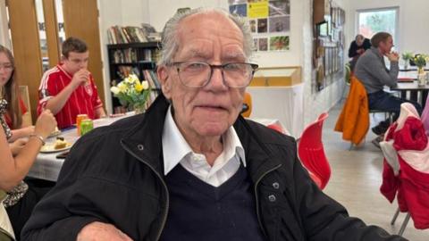 Image of Dennis Smedley - he is wearing glasses and is wearing a white shirt, navy jumper and a black coat inside a visitor centre with other people around him sat in red chairs. 
