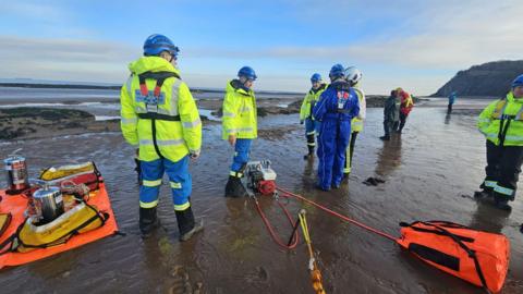 Several coastguard workers all stood on a muddy beach with the sea out. They are using a winching device and all in high-viz jackets