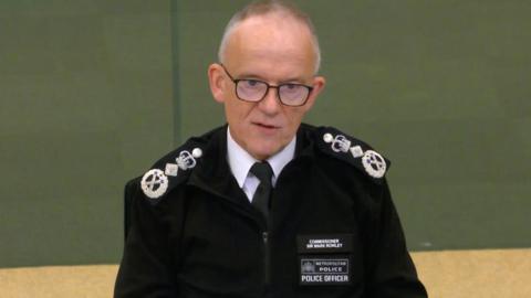 Sir Mark Rowley in black police uniform. He is sitting behind a desk at City Hall and has short grey hair and glasses.