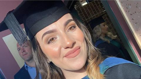 Sophie Morgan wears a black university cap and gown and smiles at the camera. She has brown short hair and brown eyes. 