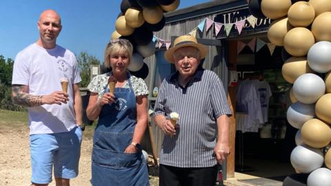 Three generations of a family - a younger man, a middle-aged woman and and elderly man - stand in front of an ice cream shop holding cones. Behind them the shop is heavily decorated with a balloon arch and bunting. 