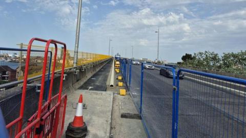 Roadworks on the Itchen Bridge in Southampton with blue and red railings either side of the footpath on the left and cars travelling along the road on the right.