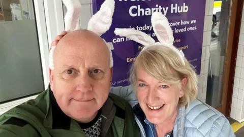 A man and a woman stand outside of a charity hub wearing white bunny ears on their heads.