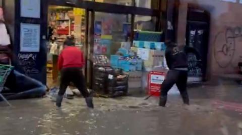 A social media image of two shop workers are seen trying to clear flood waters from outside their store