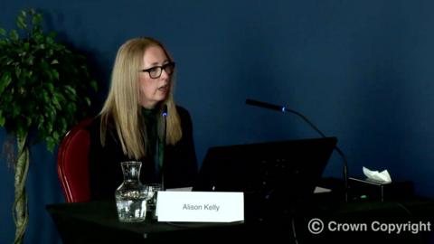 Alison Kelly, who has shoulder length reddish blonde hair and wears dark rimmed glasses and a green top under a black jacket, sits at a desk with a computer screen, microphone and a notice with her name on 