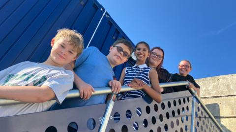Five young people stand on some stairs in front of a blue shipping container unit.