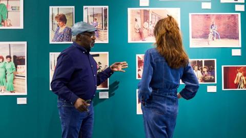 Two people dressed in denim stand in front of a blue wall featuring a number of frame photos