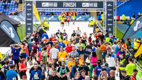 Dozens of runners gather after crossing the finish line. A clock above the finish line displays the time 2 hours, 12 minutes and 37 seconds. In the foreground a marker flag say 2 hours and two runners in navy blue t-shirts take a selfie 