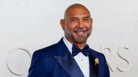 Dave Bautista smiling and looking at the camera on a red carpet. He's wearing a tux with a white shirt, blue bow tie and blue jacket. He's a bald man with a darker beard that's going grey down the sides.