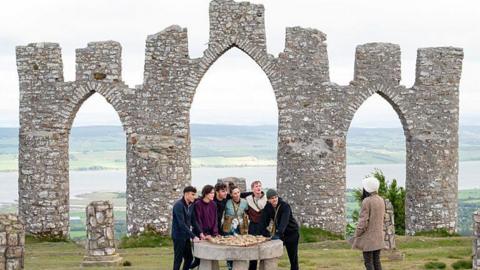A large stone folly built overlooking a body of water, a group of people are huddled below it.