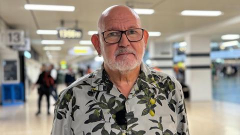 A man in a grey shirt with a lemon tree print stands in a train station. He wears glasses and a small hoop earring in one ear. 