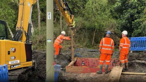 Large hole with workers in orange standing at its edge and one worker in the hole - a yellow digger is to the left.