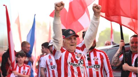 A Sunderland fan in a cap holds both arms up in celebration among other supporters holding flags