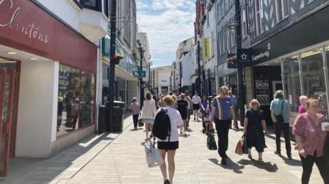 People with shopping bags walking through the high street in Douglas with shops on either side of a paved area.