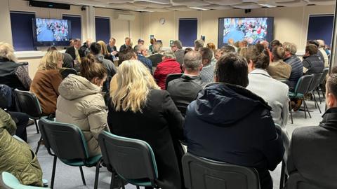 A crowd of people sitting on chairs at a council meeting. They are all facing the front, the backs of their heads are visible. Many of the attendees are wearing coats. There are two screens on the wall.
