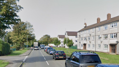 Cars line the right hand lane on a street outside a three-storey block of ageing flats in a residential area. In the left lane, a car and a lorry can be seen driving into the distance. There are trees on the grass verges opposite the flats.