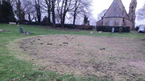 A large circle of trampled and dead grass in the middle of a graveyard. The grass is yellowed and thinned. Headstones can be seen around the edges and a church in the background.