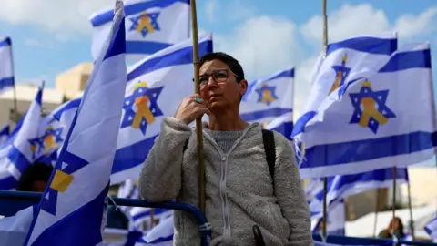 A woman looks into the distance as she stands between Israeli flags in Tel Aviv