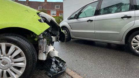 A close up image of significant damage to the front bumper of a neon green Renault car which is parked very close to a tight section of road being used by cars and vans with houses in the background. 