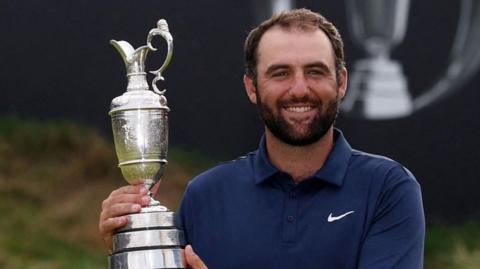 A man in a navy blue polo shirt, white trousers and a silver wrist watch holds up a trophy up and smiles at the camera.