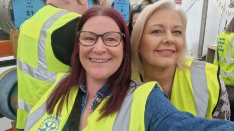 Two women wearing fluorescent Rotary Club jackets look at the camera smiling, with beer barrels behind them.