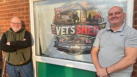 Paul Unsworth (left) and Gary Smith standing outside the Vet's Shed centre in Fleetwood. They are standing either side of a window with a banner advertising the centre. Paul has a short white beard and glasses, and is wearing a green fleece with black sleeves and jeans, and Gary is clean shaven an in a grey polo shirt Both are smiling at the camera