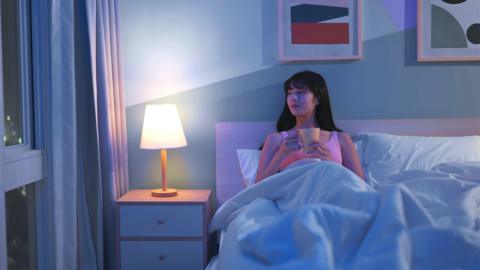 A woman is sat up in bed in an apartment drinking from a mug. She glances towards the window where the curtains are open.