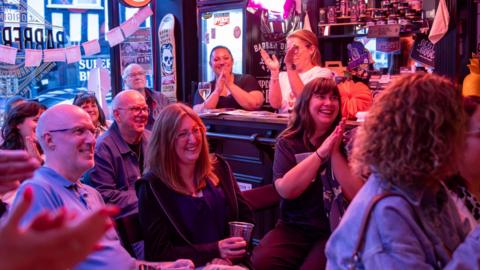 A bar with pink lighting is filled with people sat down and laughing. The people watching are clapping and holding drinks. Two women are also seen standing behind a nearby bar. 
