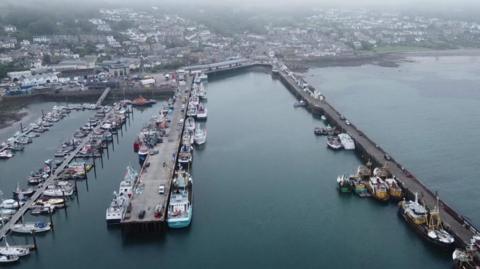 A drone shot of a harbour full of boats on a misty day. The water is a dark blue colour and there is mist and fog covering homes and the nearby town on the main land. 