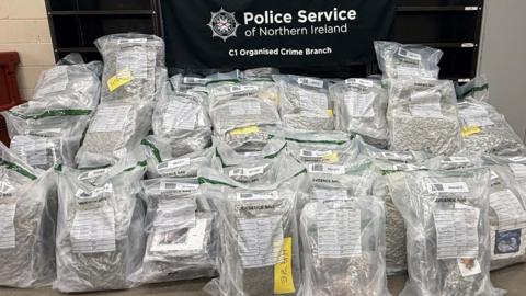 Blocks of green material can be seen inside over a dozen rucksack-sized plastic bags, which are lined and piled up in front of a banner reading "Police Service of Northern Ireland C1 Organised Crime Branch". 